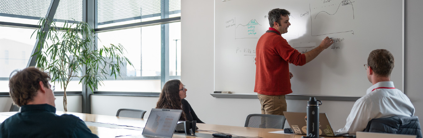 An engineer writes an equation on a whiteboard while two other engineers look on.